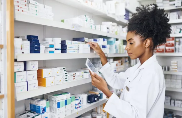 A pharmacist wearing a white lab coat is standing in front of shelves filled with various medication boxes and bottles. 