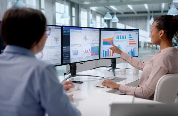 Two employees review multiple computer monitors displaying data dashboards with charts and graphs, with one person pointing to a visual trend during a discussion in a modern office.