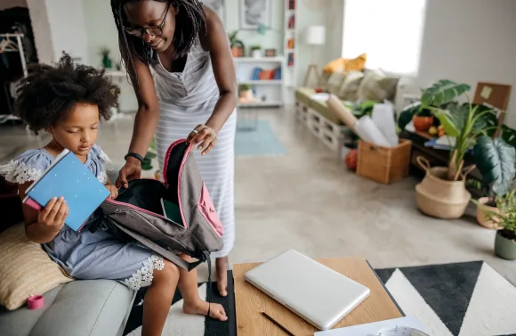 A child sits on a couch holding notebooks while packing a backpack with assistance from an adult in a bright living room. A laptop and notebook rest on a nearby coffee table, suggesting preparation for school or learning activities at home.