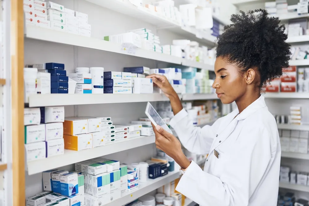 A pharmacist wearing a white lab coat is standing in front of shelves filled with various medication boxes and bottles. 