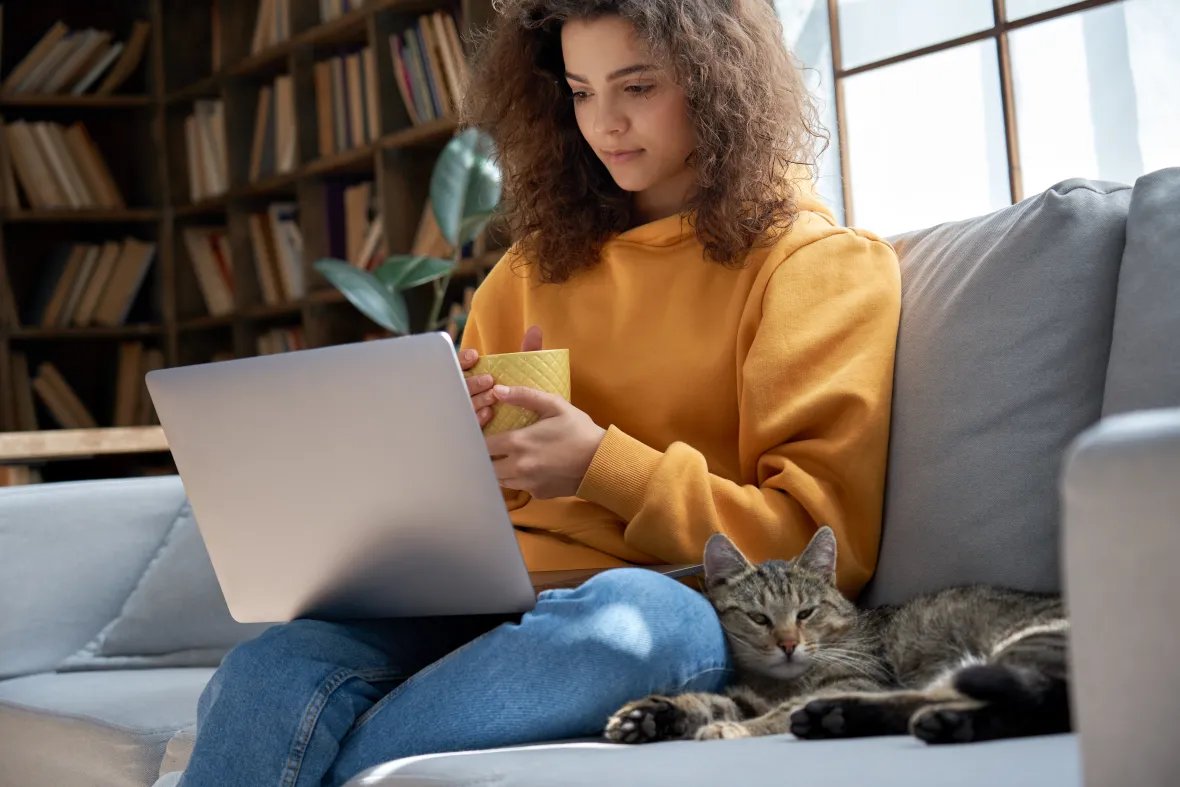 Person with laptop and mug on couch, cat beside them, bookshelves and window in background.