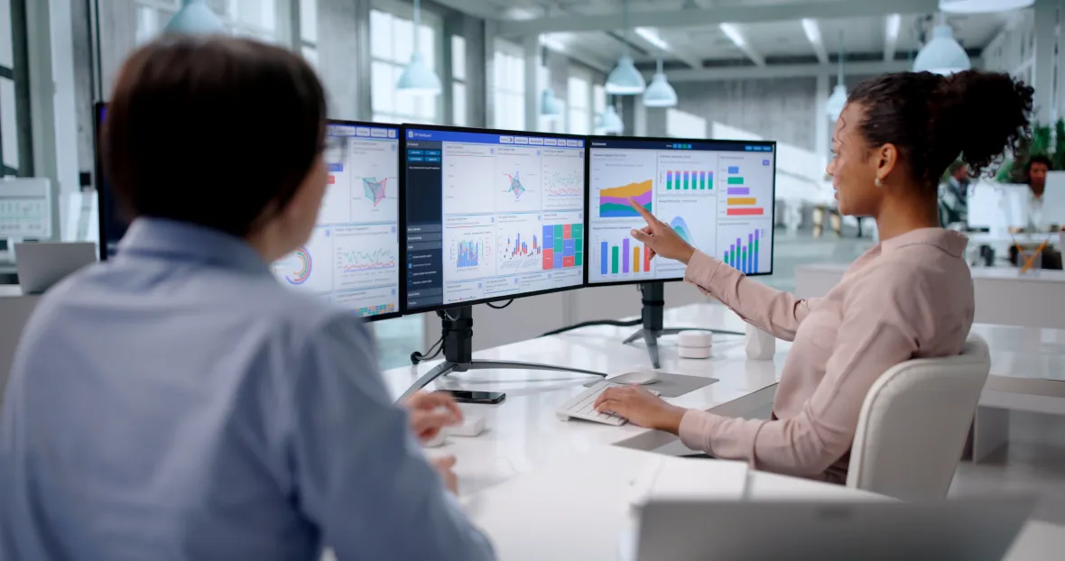 Two employees review multiple computer monitors displaying data dashboards with charts and graphs, with one person pointing to a visual trend during a discussion in a modern office.