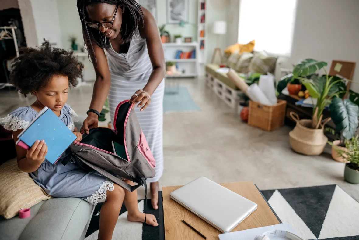 A child sits on a couch holding notebooks while packing a backpack with assistance from an adult in a bright living room. A laptop and notebook rest on a nearby coffee table, suggesting preparation for school or learning activities at home.
